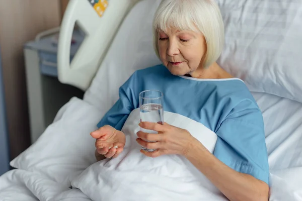 Femme âgée couchée au lit et tenant des pilules avec un verre d'eau à l'hôpital — Photo de stock