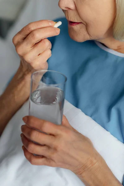 Ausgeschnittene Ansicht einer Seniorin, die im Bett liegt, ein Glas Wasser in der Hand hält und im Krankenhaus Tabletten nimmt — Stockfoto