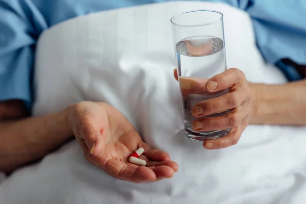 Selektiver Fokus der Seniorin, die im Bett liegt und Tabletten mit Wasserglas im Krankenhaus hält — Stockfoto