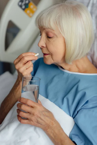 Femme âgée couchée au lit, tenant un verre d'eau et prenant une pilule à l'hôpital — Photo de stock