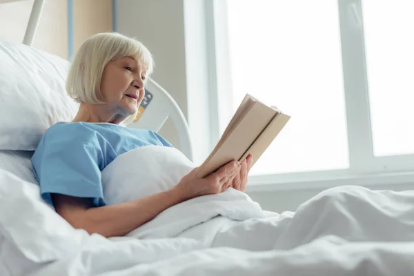 Senior woman with grey hair lying in bed and reading book in hospital — Stock Photo