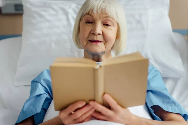 Mujer mayor acostada en la cama y leyendo libro en el hospital - foto de stock