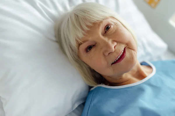 Selektiver Fokus einer lächelnden Seniorin mit grauen Haaren, die im Krankenhaus im Bett liegt — Stockfoto