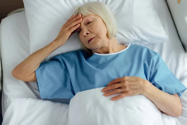 Bouleversé femme âgée couchée dans le lit d'hôpital, toucher le front et avoir mal à la tête — Photo de stock