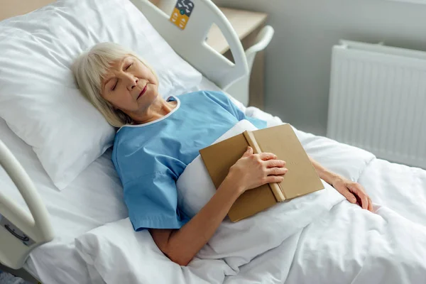 Enfoque selectivo de la mujer mayor con libro durmiendo en la cama en el hospital - foto de stock