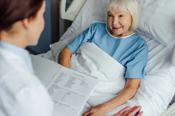 Arzt hält Diagnose und berät lächelnde Seniorin im Krankenhausbett — Stockfoto