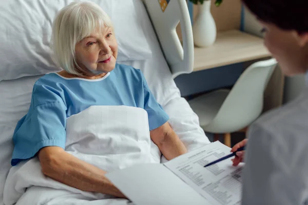 Лікар тримає діагноз і консультує старшу жінку з сірим волоссям, лежачи в лікарняному ліжку — Stock Photo