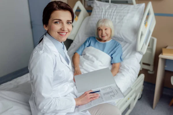 Smiling female doctor sitting on bed with diagnosis and looking at camera while senior woman lying in bed in hospital — Stock Photo