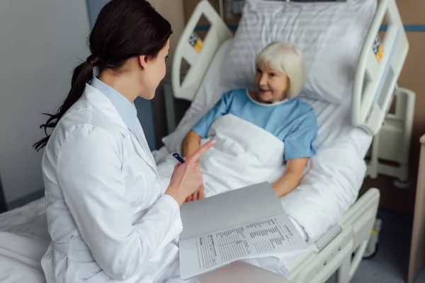 Ärztin sitzt auf Bett, hält Diagnose und zeigt mit Finger, während Seniorin im Krankenhaus im Bett liegt — Stockfoto