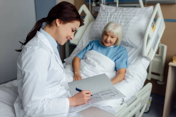 Sonriente doctora sentada en la cama y escribiendo diagnóstico mientras mujer mayor acostada en la cama en el hospital - foto de stock