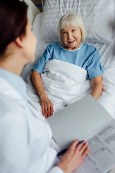 Femme médecin assis sur le lit et tenant le diagnostic tandis que la femme âgée couchée dans le lit à l'hôpital — Photo de stock