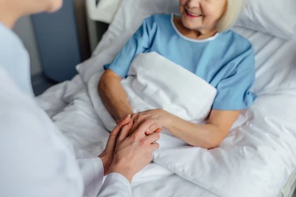 Visão cortada do médico feminino de mãos dadas com a mulher idosa deitada na cama no hospital — Fotografia de Stock