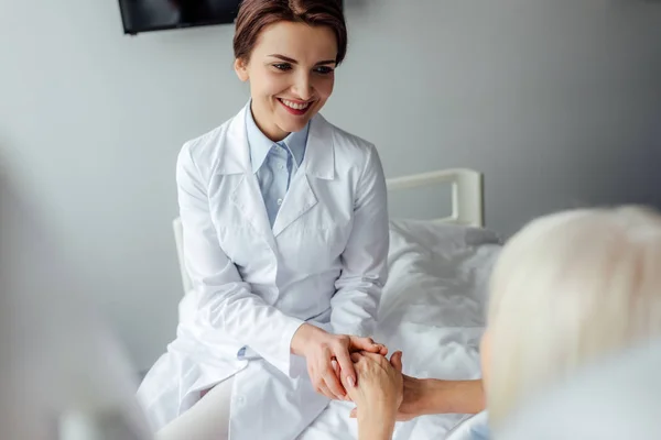 Sorridente medico femminile che si tiene per mano con la donna anziana sdraiata a letto in ospedale — Foto stock