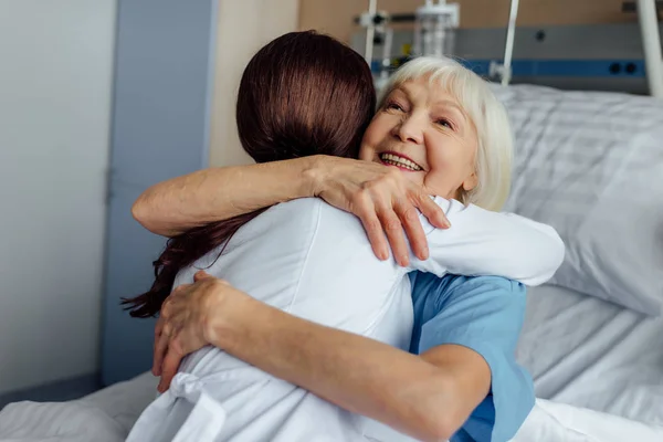 Mulher sênior feliz deitado na cama e abraçando médico feminino no hospital — Fotografia de Stock