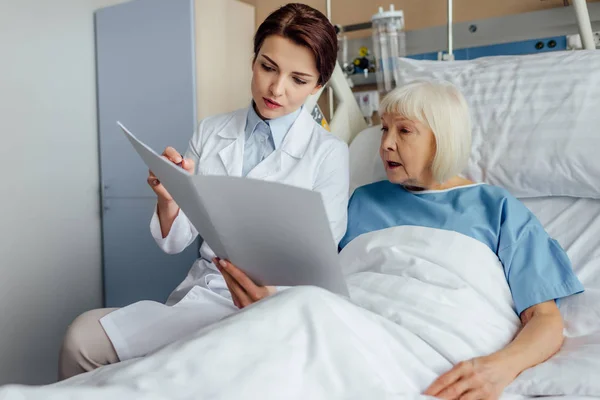 Серйозна жінка-лікар, яка тримає діагноз і консультує здивовану старшу жінку, що лежить в лікарняному ліжку — стокове фото