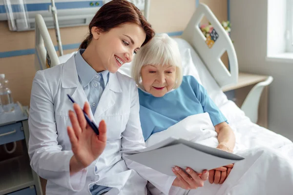 Happy female doctor holding diagnosis and consulting senior woman lying in hospital bed — Stock Photo