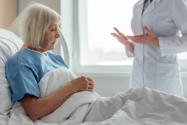 Médico mujer consultando triste mujer mayor con el pelo gris acostado en la cama del hospital - foto de stock
