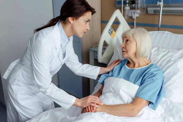 Medico femminile che si tiene per mano e consulenza triste donna anziana sdraiata nel letto d'ospedale — Foto stock