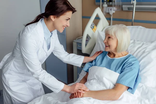 Feliz médico femenino cogido de la mano y consultando a la mujer mayor acostada en la cama del hospital - foto de stock