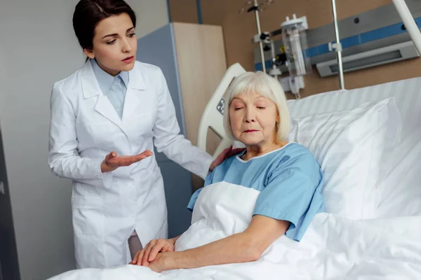 Female doctor holding hands and consulting upset senior woman lying in hospital bed — Stock Photo