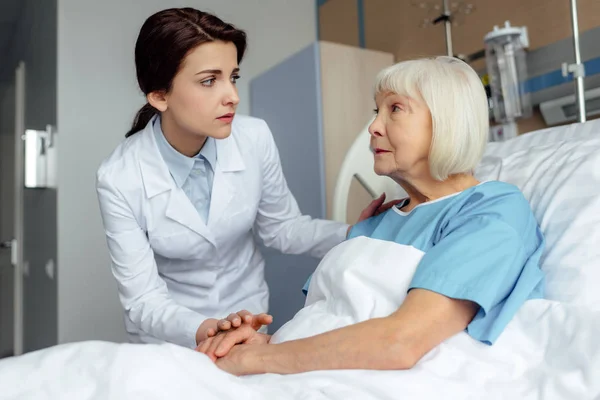 Preoccupato medico femminile tenendosi per mano e consultando la donna anziana sdraiata nel letto d'ospedale — Foto stock