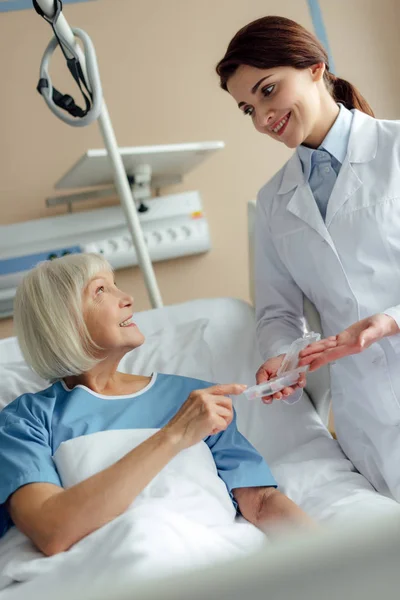 Sonriente médico mujer dando pastillas a la mujer mayor acostada en la cama del hospital - foto de stock