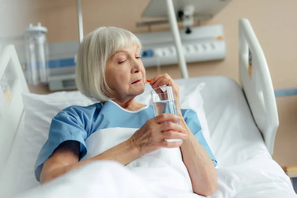 Selective focus of senior woman lying in bed, holding glass of water and taking pill in hospital — Stock Photo