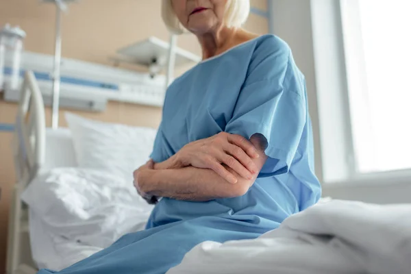 Cropped view of senior woman with arms crossed sitting on bed in hospital ward — Stock Photo