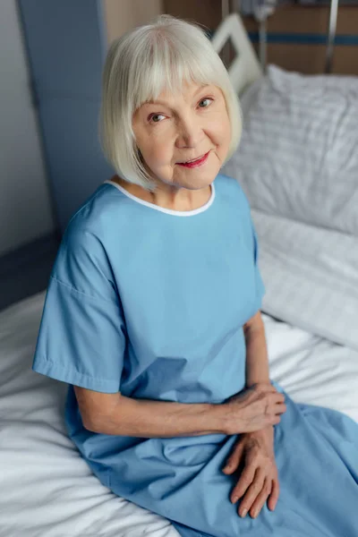 Mujer mayor sonriente con las manos dobladas sentadas en la cama y mirando a la cámara en el hospital - foto de stock