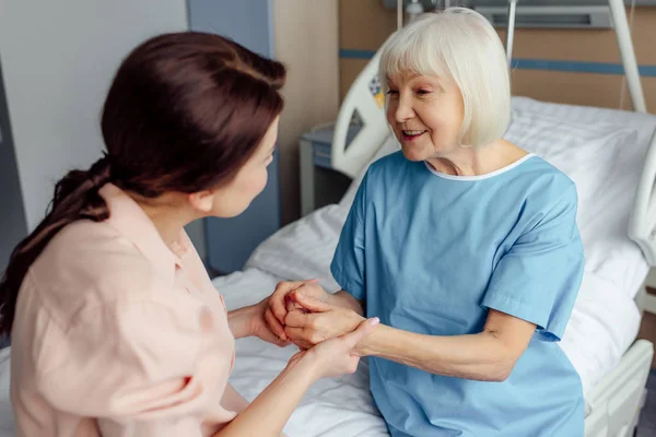 Glückliche Seniorin und Tochter auf Bett sitzend und Händchen haltend im Krankenhaus — Stockfoto