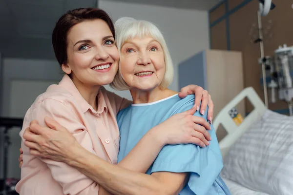 Glückliche Seniorin und Tochter sitzen auf Bett und umarmen sich im Krankenhaus — Stockfoto