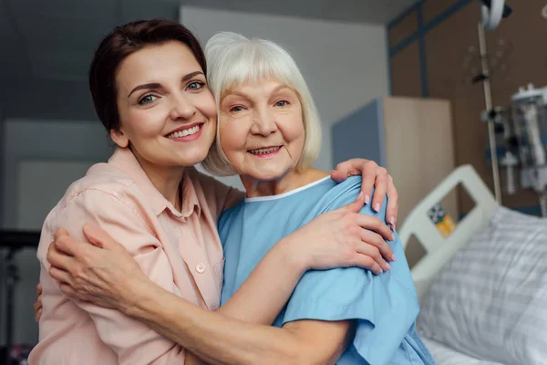 Souriant femme âgée et fille assise sur le lit, regardant la caméra et embrassant à l'hôpital — Photo de stock