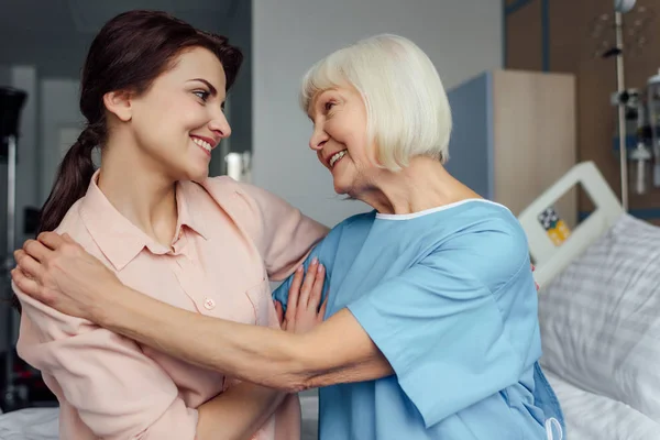 Heureuse femme âgée et fille assise sur le lit et embrassant à l'hôpital — Photo de stock