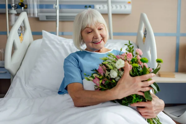 Smiling recovering senior woman lying in bed with flowers in hospital — Stock Photo
