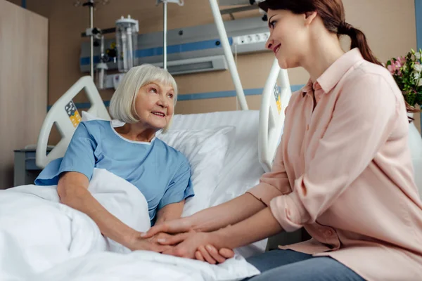 Seniorin und Tochter sitzen auf Bett, schauen einander an und halten sich im Krankenhaus an den Händen — Stockfoto
