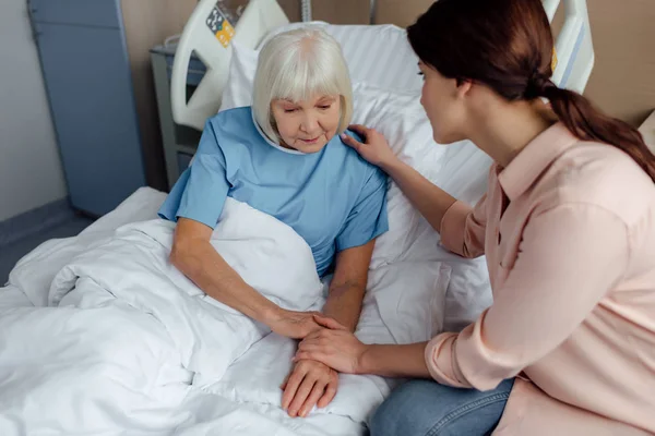 Дочь сидит рядом со старшей матерью на кровати и держит руки в больнице — стоковое фото