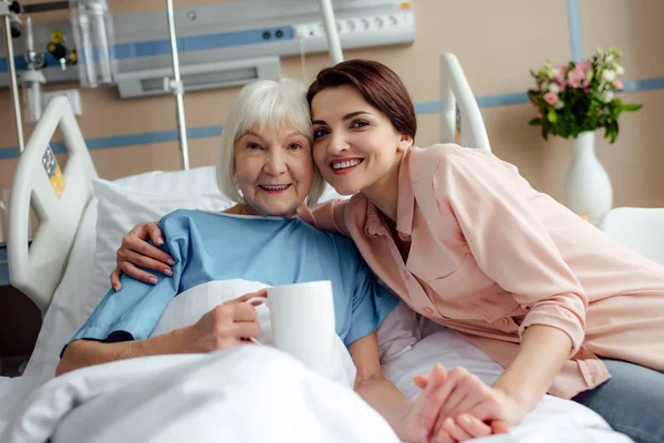 Пожилая женщина и дочь с чашкой чая смотрят в камеру и обнимаются на кровати в больнице — стоковое фото