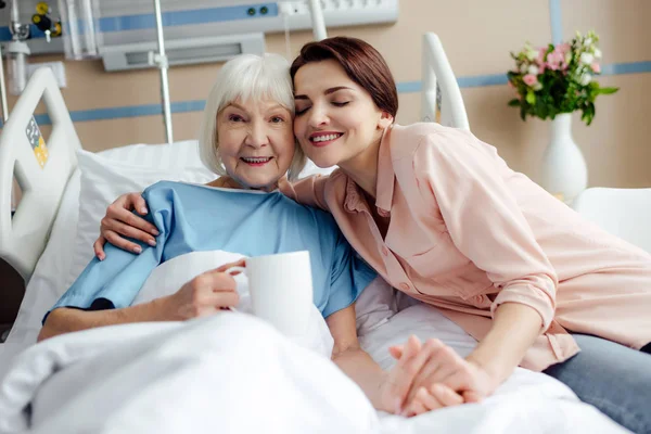 Счастливая пожилая женщина и дочь с чашкой чая обнимаются на кровати в больнице — стоковое фото