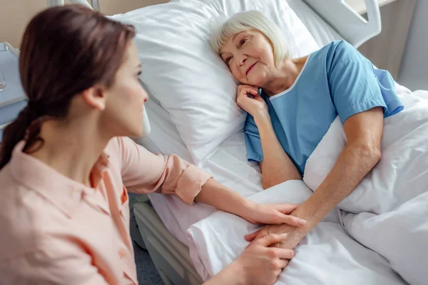 Hija sentada cerca de la madre mayor en la cama y cogida de la mano en el hospital - foto de stock