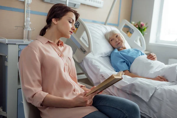 Красивая дочь читает книгу больной старшей матери, лежащей в постели в больнице — стоковое фото