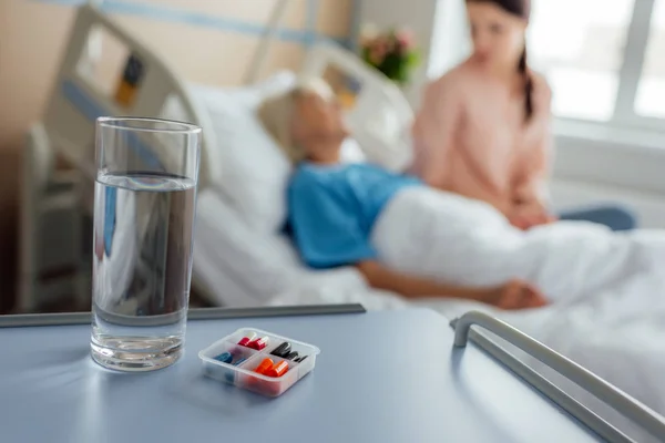 Foco seletivo de vidro de água e medicina com paciente e visitante em segundo plano — Fotografia de Stock