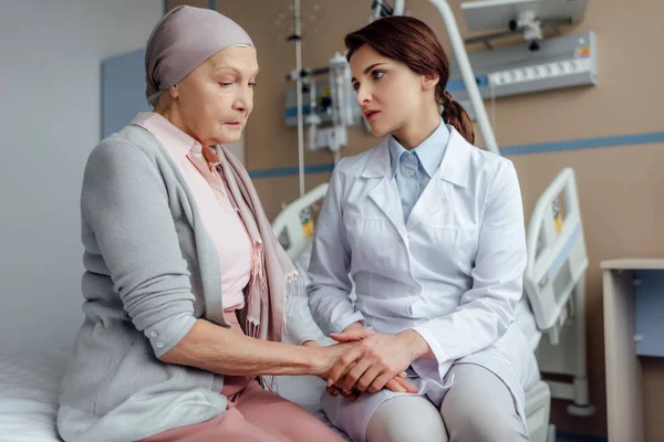 Doctora preocupada cogida de la mano con una mujer mayor en pañuelo con cáncer en el hospital — Stock Photo
