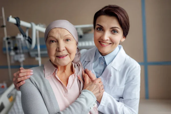 Feliz médico femenino abrazando a la mujer mayor en el pañuelo con cáncer y mirando a la cámara en el hospital — Stock Photo