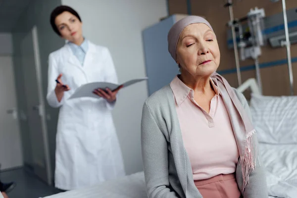 Triste donna anziana in fazzoletto con cancro seduta sul letto d'ospedale con medico donna che tiene la diagnosi sullo sfondo — Foto stock