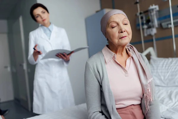 Sad senior woman in kerchief with cancer sitting on hospital bed with female doctor holding diagnosis on background — Stock Photo