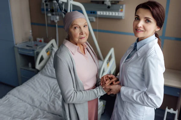 Medico donna che guarda la macchina fotografica e si tiene per mano con la donna anziana in fazzoletto con cancro in ospedale — Foto stock