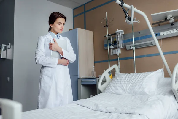 Sad female doctor with diagnosis looking at empty bed in hospital ward — Stock Photo