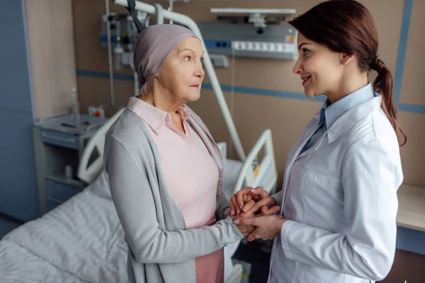 Smiling female doctor holding hands with senior woman in kerchief with cancer in hospital — Stock Photo