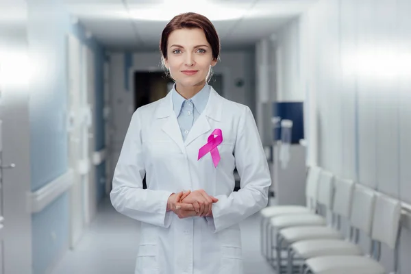 Female doctor with pink ribbon and folded hands looking at camera in hospital, breast cancer awareness concept — Stock Photo