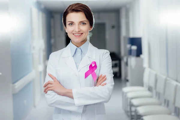 Smiling female doctor with pink ribbon and arms crossed in hospital, breast cancer awareness concept — Stock Photo